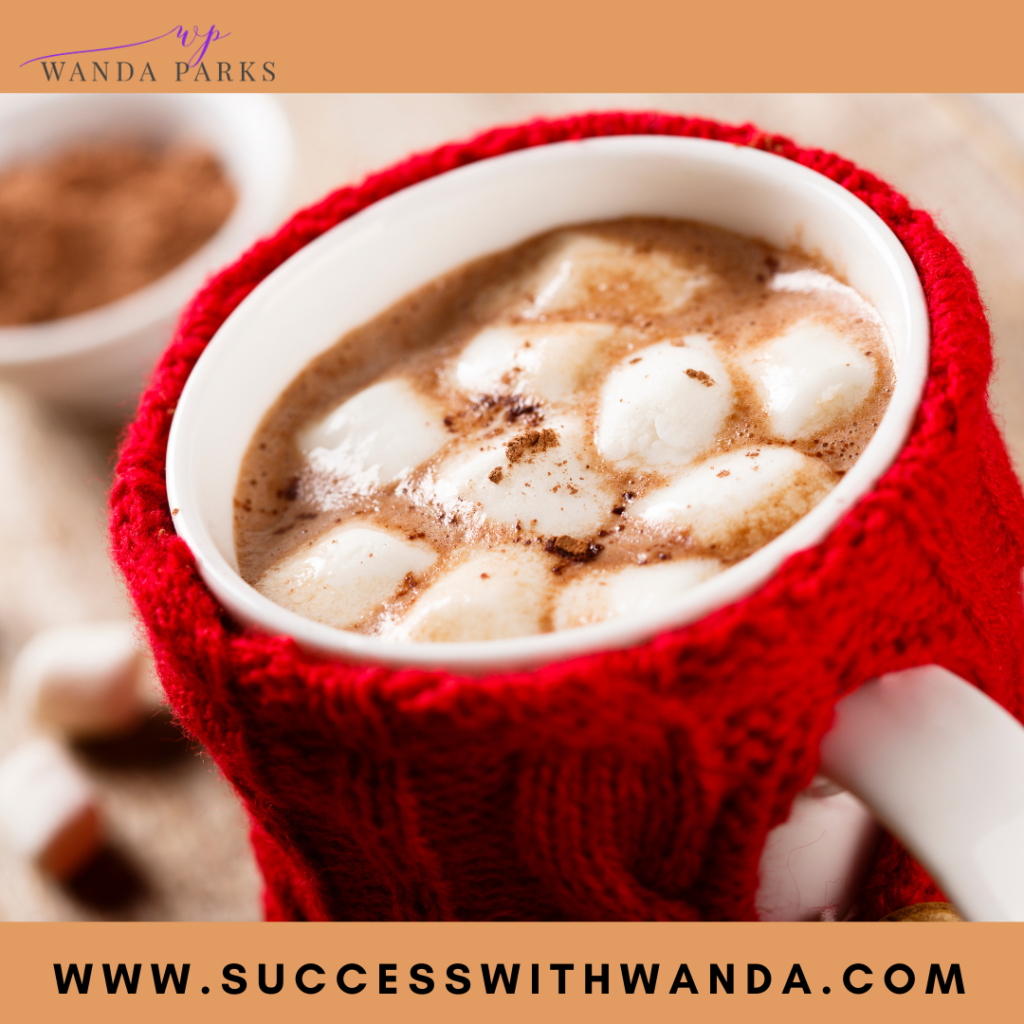 white coffee cup enclosed in a red knit cup holder, filled with hot cocoa and multiple pieces of white marshmallows

